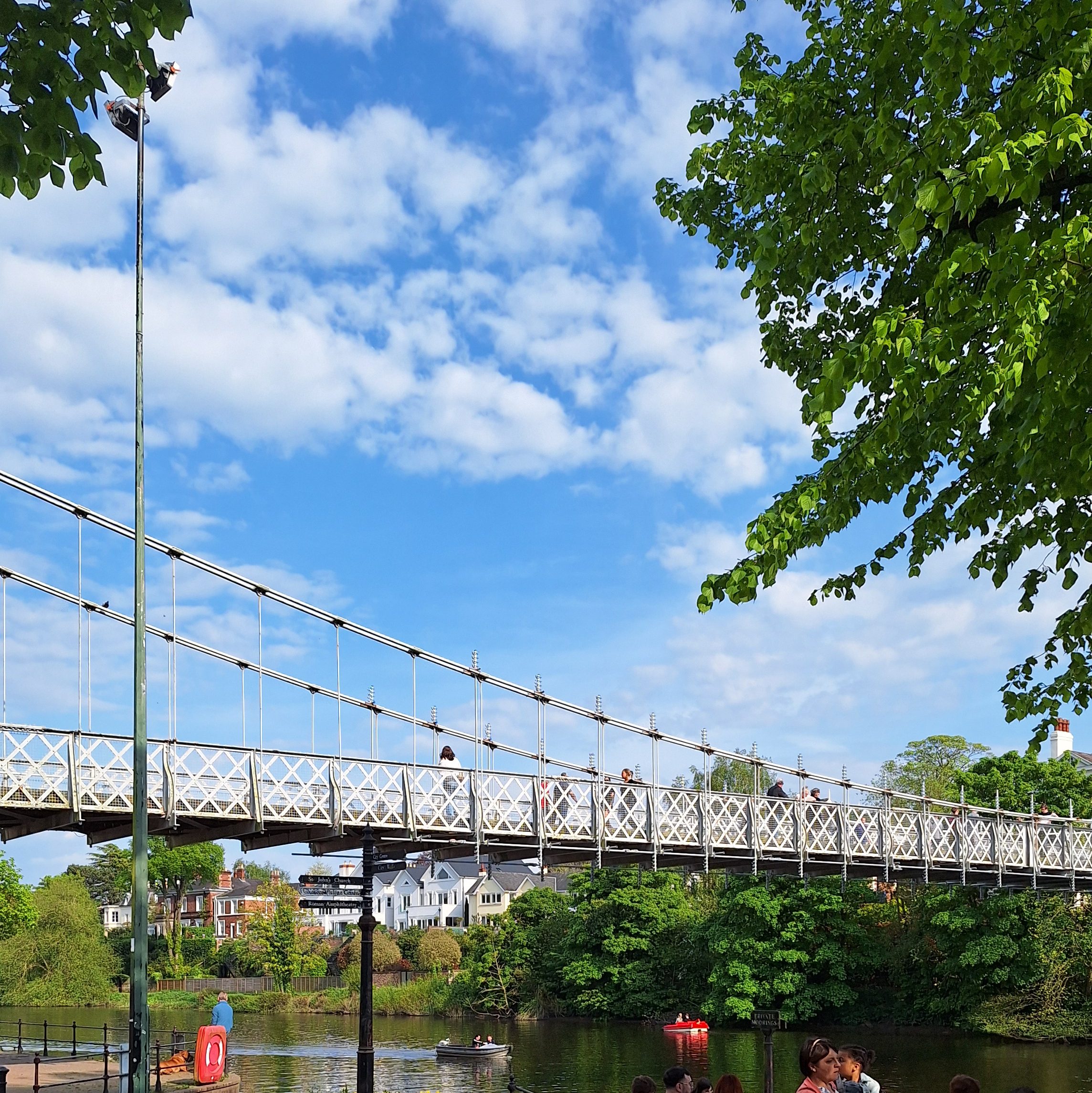 A view of a bridge over the River Dee in Chester, There is a boat on the water and people in the river bank.