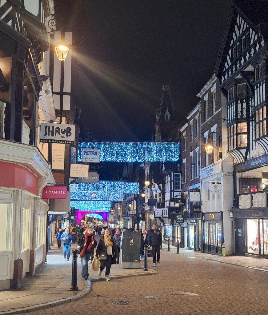 A view of Eastgate street in Chester. Christmas lights illuminate the street. Shoppers are carrying bags.