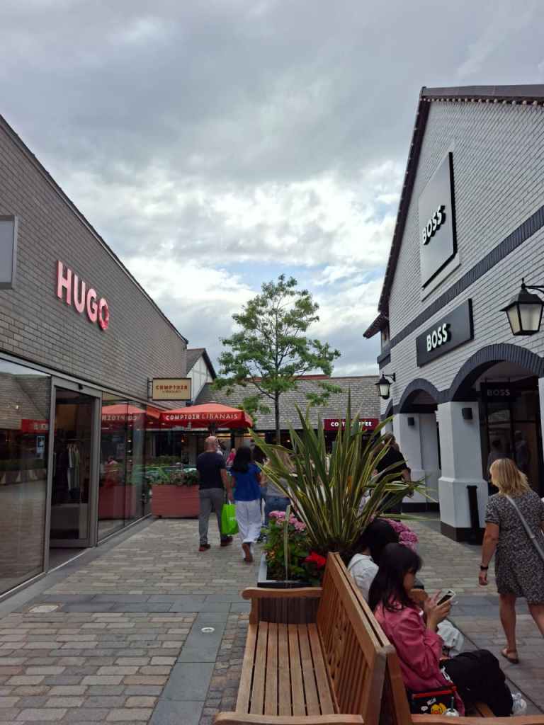 People shopping at Cheshire Oaks Outlet village. Some people are also sitting outside on a dull day on benches.